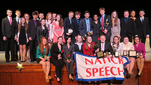 Picture of Luke with a large team of students in professional attire holding a banner that reads NATICK SPEECH